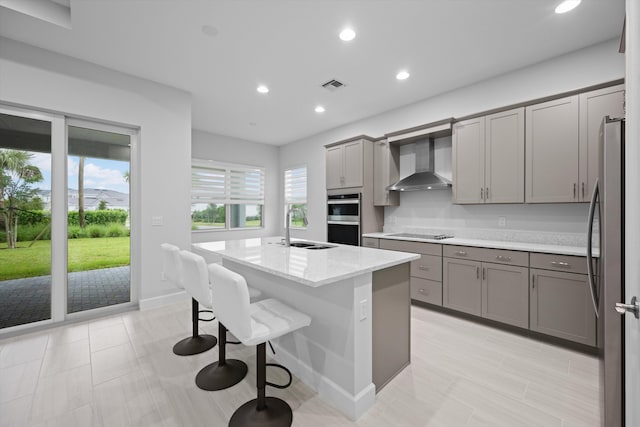 kitchen with an island with sink, wall chimney exhaust hood, sink, appliances with stainless steel finishes, and gray cabinets