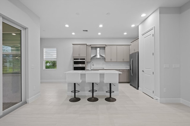 kitchen featuring wall chimney range hood, gray cabinets, a breakfast bar, a center island with sink, and stainless steel appliances