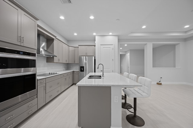 kitchen with wall chimney range hood, gray cabinets, a breakfast bar, a center island with sink, and sink