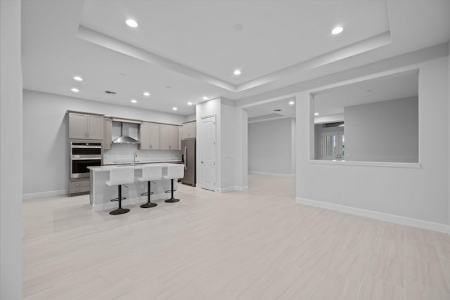 kitchen with wall chimney range hood, gray cabinets, a kitchen island with sink, a kitchen breakfast bar, and stainless steel appliances