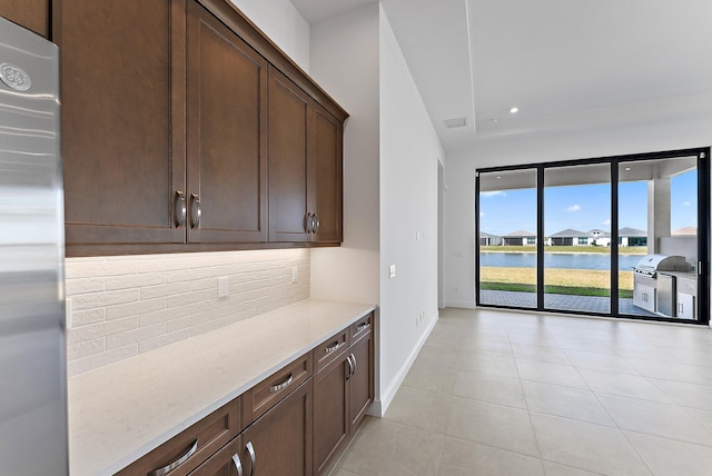 bar featuring stainless steel fridge, a water view, dark brown cabinetry, light stone countertops, and decorative backsplash