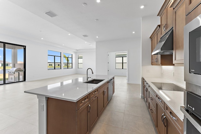 kitchen featuring sink, appliances with stainless steel finishes, tasteful backsplash, light stone countertops, and an island with sink