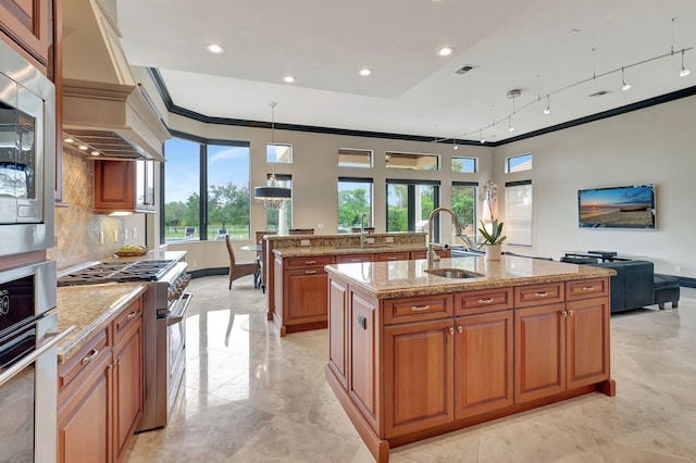 kitchen featuring stainless steel appliances, decorative light fixtures, light stone counters, custom range hood, and a center island with sink