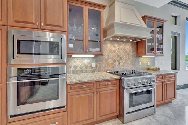 kitchen with stainless steel appliances, premium range hood, light stone countertops, and backsplash