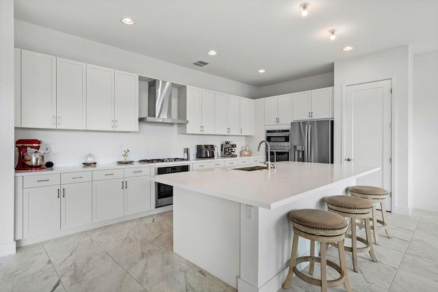 kitchen with wall chimney range hood, a kitchen island with sink, sink, white cabinets, and appliances with stainless steel finishes