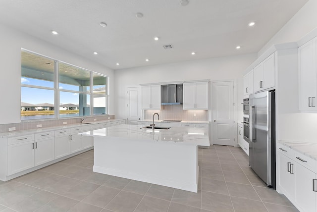 kitchen featuring an island with sink, high quality fridge, wall chimney range hood, and light stone counters