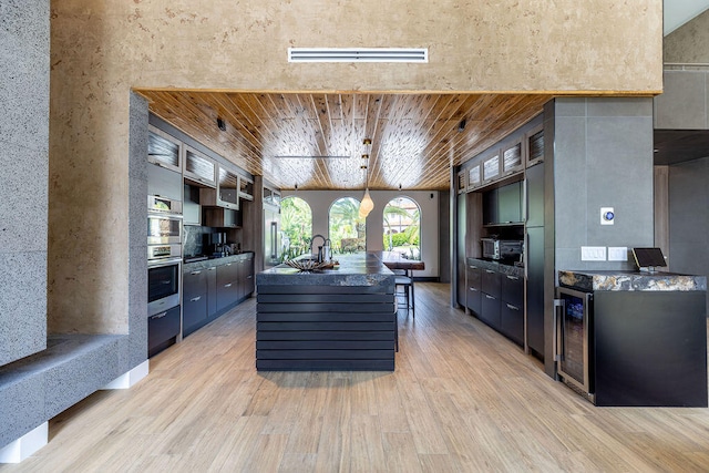 kitchen featuring hanging light fixtures, wooden ceiling, wine cooler, an island with sink, and light hardwood / wood-style floors