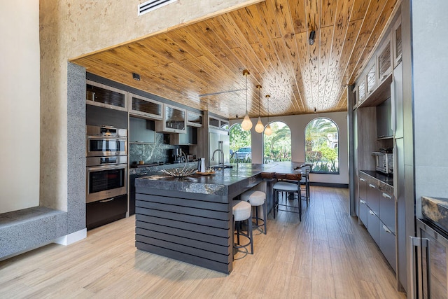 kitchen with wooden ceiling, double oven, decorative light fixtures, a center island with sink, and light wood-type flooring