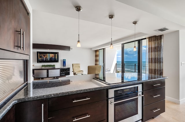 kitchen with cooktop, oven, hanging light fixtures, dark brown cabinetry, and dark stone counters