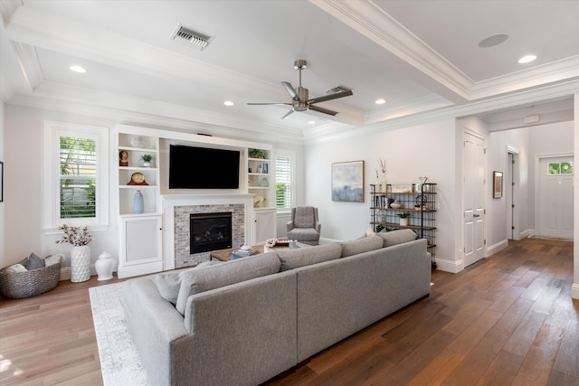 living room featuring ceiling fan, wood-type flooring, beamed ceiling, a stone fireplace, and crown molding