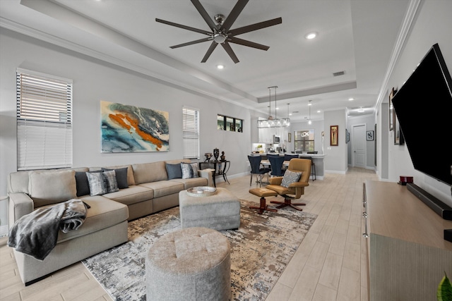 living room with ceiling fan with notable chandelier, ornamental molding, light wood-type flooring, and a raised ceiling