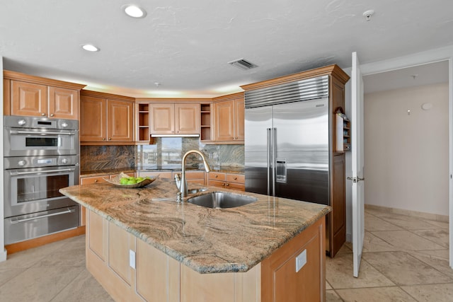 kitchen featuring a kitchen island with sink, sink, decorative backsplash, light stone counters, and stainless steel appliances