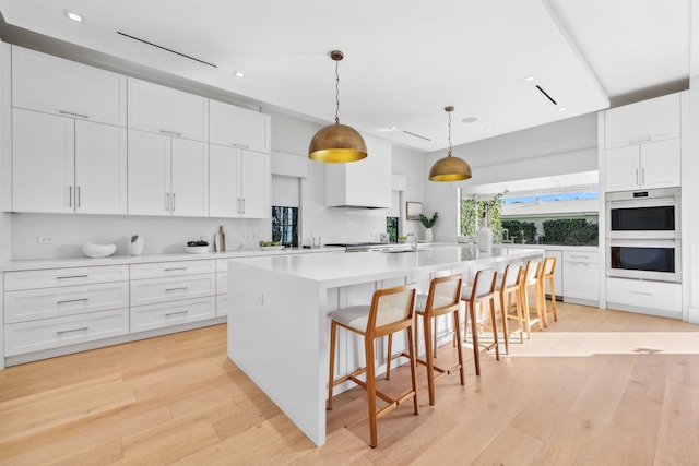 kitchen featuring white double oven, pendant lighting, light hardwood / wood-style floors, white cabinetry, and an island with sink