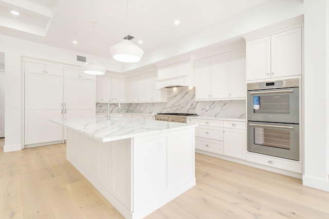 kitchen featuring light wood-style floors, decorative backsplash, an island with sink, and double oven