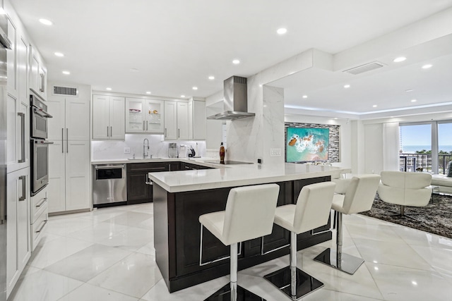 kitchen featuring a sink, wall chimney exhaust hood, light countertops, white cabinetry, and a kitchen breakfast bar