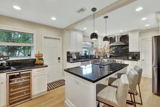 kitchen with wall chimney range hood, wine cooler, backsplash, a kitchen island, and white cabinetry