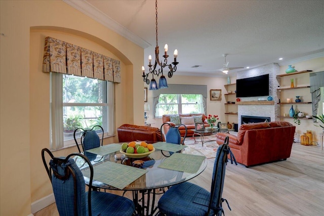 dining area featuring a stone fireplace, light hardwood / wood-style floors, ceiling fan with notable chandelier, ornamental molding, and a textured ceiling