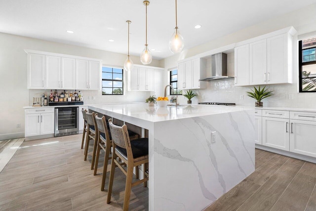 kitchen with wall chimney range hood, white cabinetry, wine cooler, and a large island with sink