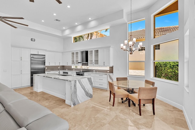 kitchen featuring wall chimney range hood, light stone countertops, appliances with stainless steel finishes, white cabinetry, and a high ceiling