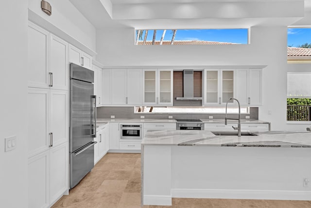 kitchen featuring stainless steel built in fridge, wall chimney exhaust hood, sink, white cabinetry, and light stone counters