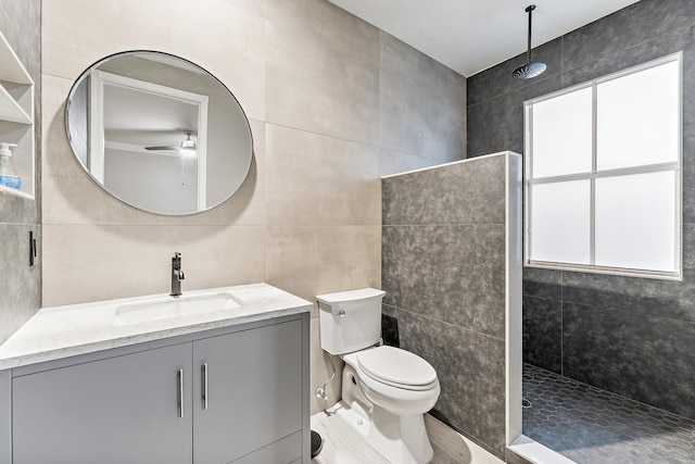 bathroom with ceiling fan, toilet, tiled shower, vanity, and tile walls