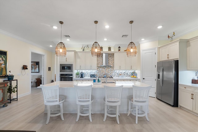 kitchen featuring wall chimney range hood, hanging light fixtures, a center island with sink, appliances with stainless steel finishes, and light hardwood / wood-style floors