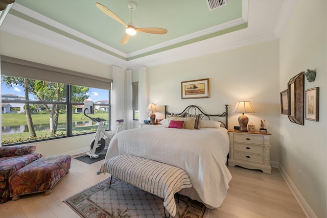 bedroom featuring ceiling fan, a tray ceiling, ornamental molding, light hardwood / wood-style floors, and a water view