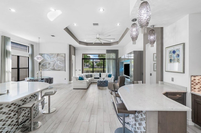 kitchen featuring a raised ceiling, light wood-type flooring, ceiling fan with notable chandelier, decorative light fixtures, and light stone counters