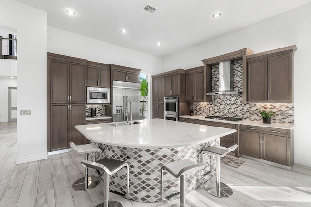 kitchen featuring light wood-type flooring, a kitchen bar, wall chimney exhaust hood, built in appliances, and a kitchen island with sink