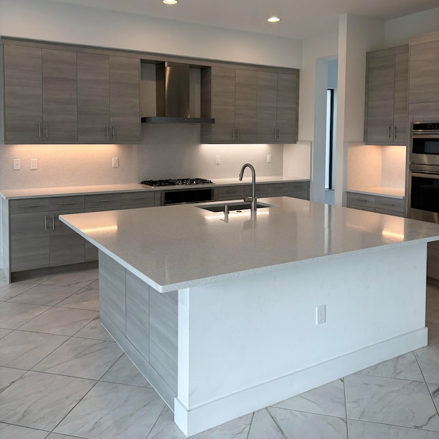 kitchen with gray cabinetry, wall chimney range hood, sink, and an island with sink