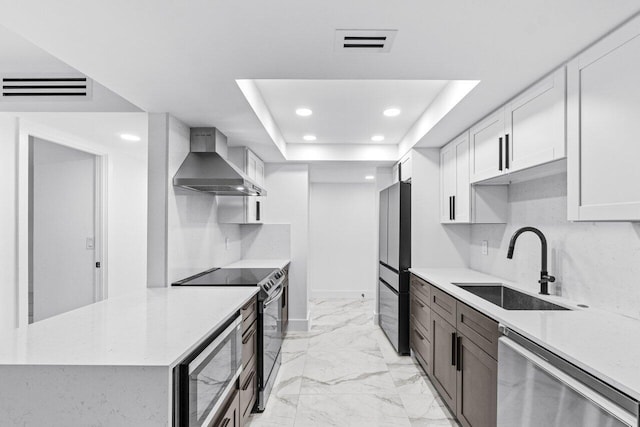 kitchen featuring wall chimney range hood, sink, a tray ceiling, stainless steel appliances, and white cabinets