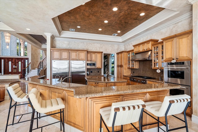 kitchen featuring a large island with sink, built in appliances, ornamental molding, a breakfast bar, and a raised ceiling