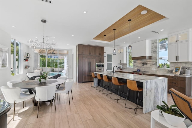 kitchen featuring an island with sink, backsplash, pendant lighting, a chandelier, and white cabinetry