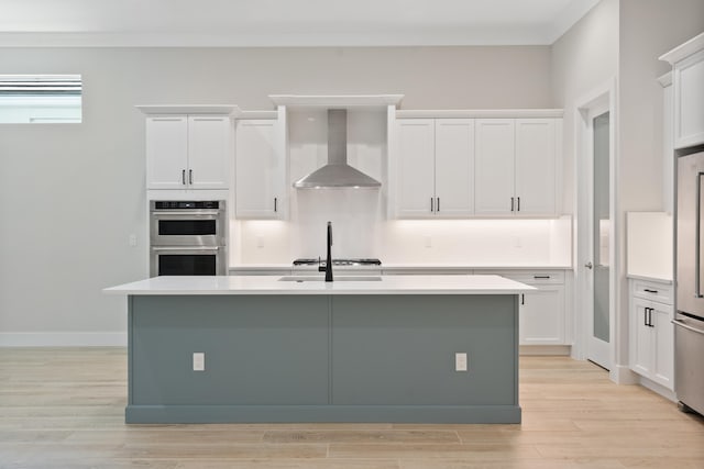 kitchen featuring wall chimney exhaust hood, an island with sink, stainless steel appliances, and light wood-type flooring