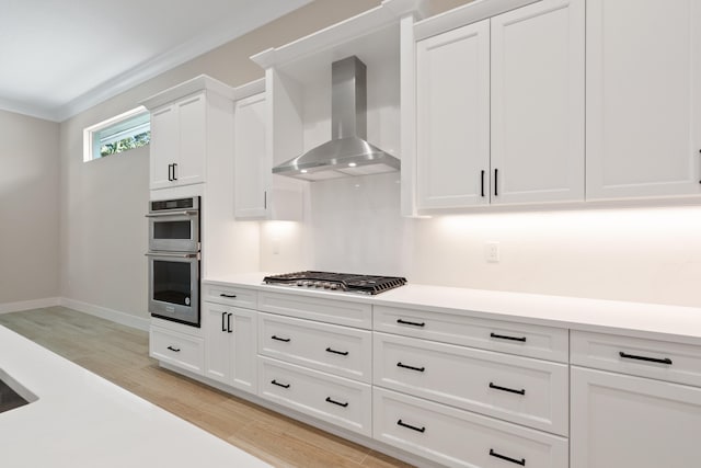 kitchen with wall chimney range hood, appliances with stainless steel finishes, light wood-type flooring, white cabinetry, and crown molding