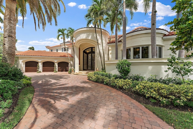 mediterranean / spanish-style house with an attached garage, a tiled roof, decorative driveway, and stucco siding
