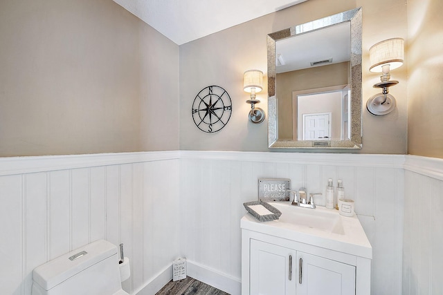 half bathroom with visible vents, a wainscoted wall, vanity, toilet, and a decorative wall