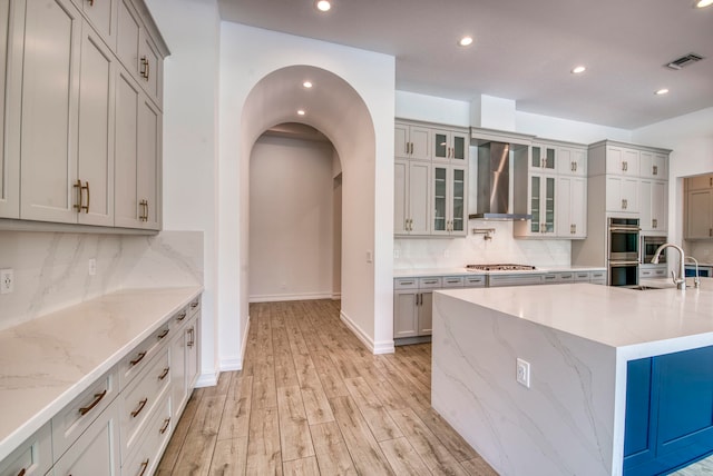 kitchen featuring wall chimney range hood, appliances with stainless steel finishes, light wood-type flooring, sink, and an island with sink