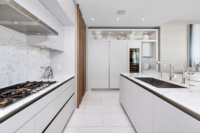 kitchen with wall chimney range hood, white cabinets, light stone countertops, and sink