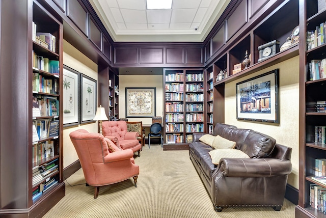 sitting room with bookshelves, a drop ceiling, and light colored carpet