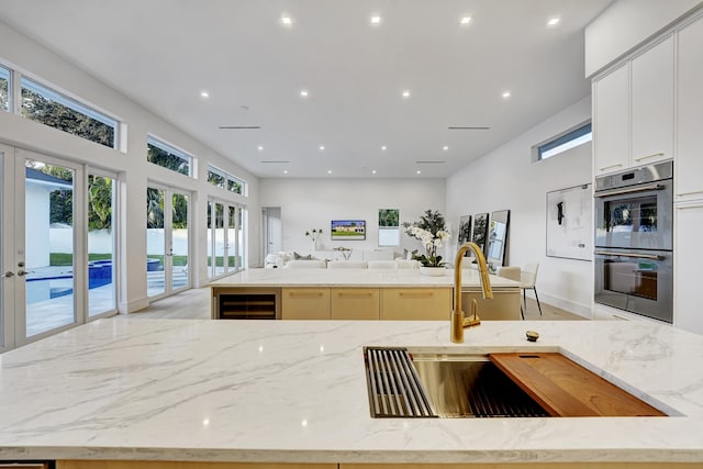 kitchen with sink, an island with sink, stainless steel double oven, wine cooler, and light stone counters