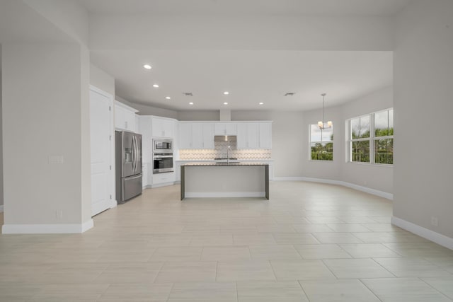 kitchen featuring appliances with stainless steel finishes, an island with sink, backsplash, white cabinets, and a notable chandelier