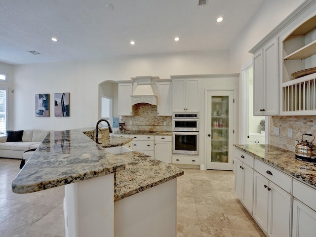 kitchen featuring premium range hood, sink, stainless steel double oven, light stone countertops, and a center island with sink