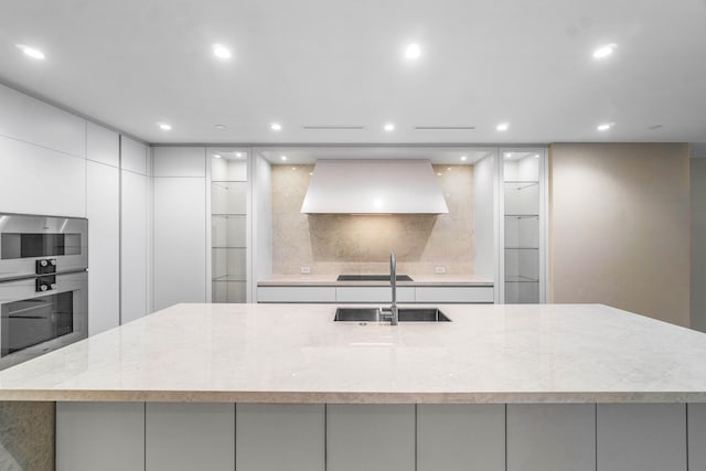 kitchen with sink, premium range hood, double oven, light stone countertops, and white cabinets