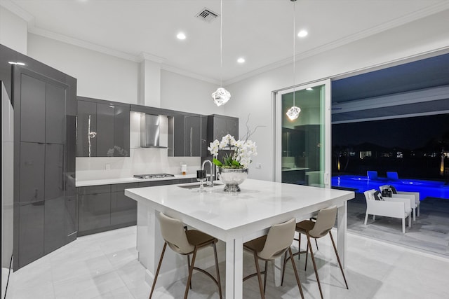 kitchen featuring wall chimney exhaust hood, a kitchen breakfast bar, an island with sink, sink, and decorative light fixtures