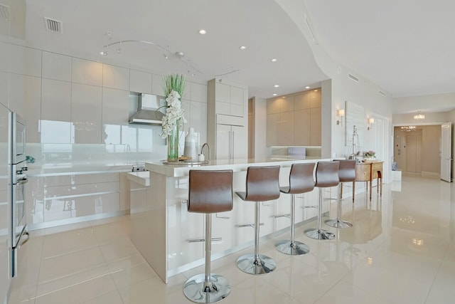 kitchen with a kitchen bar, wall chimney range hood, a center island with sink, white cabinetry, and light tile patterned flooring