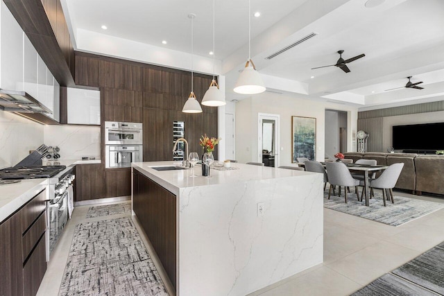 kitchen featuring stainless steel double oven, a kitchen island with sink, a tray ceiling, sink, and decorative light fixtures