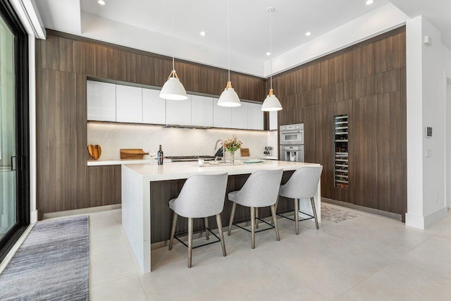 kitchen featuring stainless steel double oven, an island with sink, hanging light fixtures, backsplash, and white cabinets