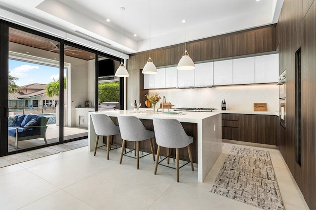 kitchen featuring stainless steel gas cooktop, backsplash, dark brown cabinetry, decorative light fixtures, and a kitchen island with sink