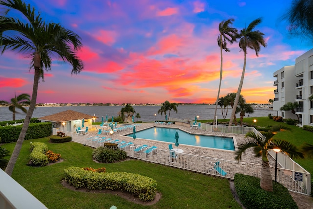 pool at dusk featuring a patio, a water view, and a lawn